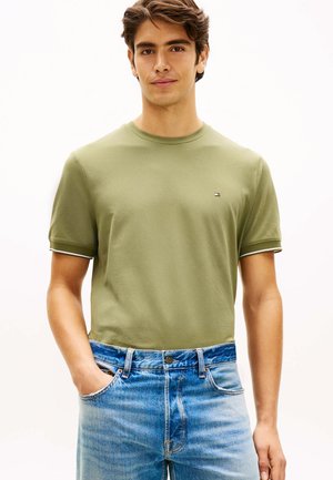 Jeune homme portant un t-shirt à manches courtes vert olive et un jean bleu clair, debout avec une main dans la poche devant un fond uni.