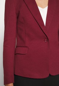 Bordeauxrode blazer met een getextureerde stof, enkele knoopsluiting en zijzakken met een getailleerde pasvorm. Witte top met V-hals eronder.