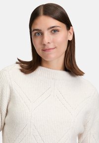 Junge Frau mit glatten, braunen Haaren, die einen weißen strukturierten Pullover trägt, mit leichtem Lächeln frontal zur Kamera blickend, grauer Hintergrund.