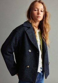 Plašči iz volnenega mešanice z dvojno zapenjanjem v temno modri barvi s teksturiranim houndstooth vzorcem, velikimi gumbi in obrezanimi lapelami. Priložnostni kroj.