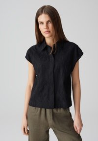 Camicia nera a maniche corte con un motivo a onde testurizzato, dotata di colletto e chiusura a bottoni, abbinata a pantaloni beige.