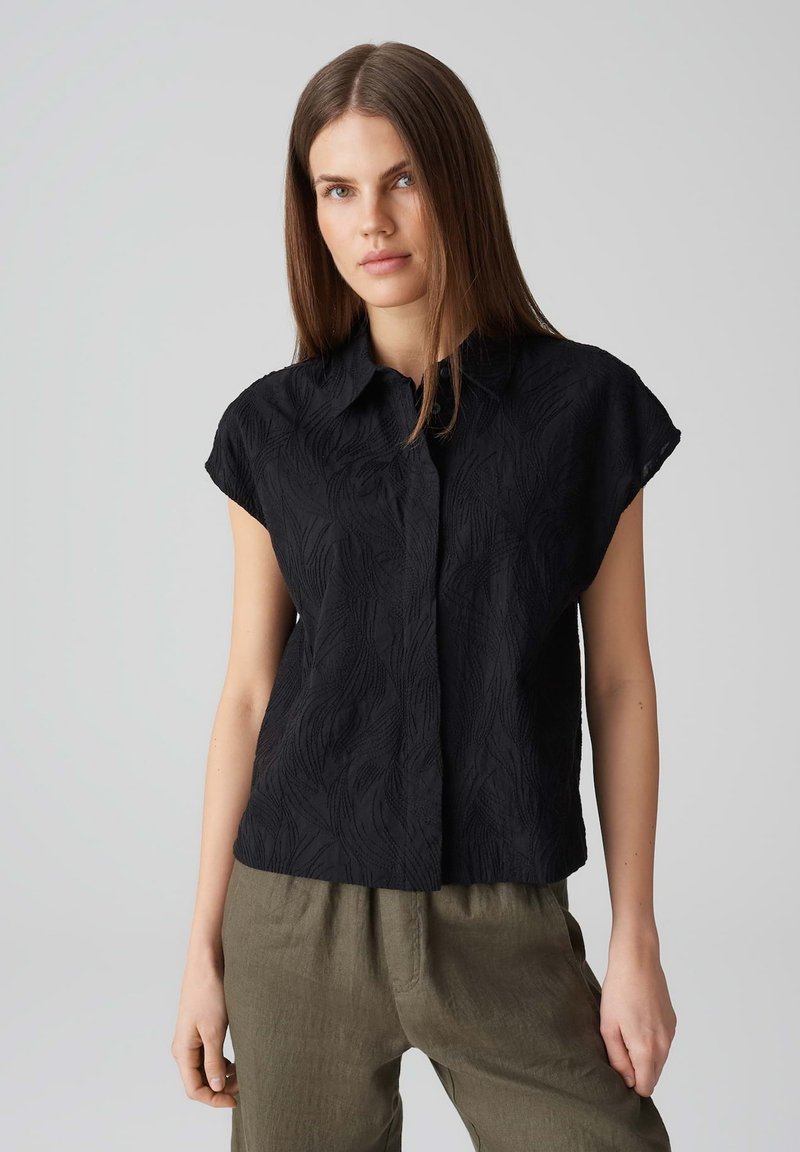 Camicia nera a maniche corte con un motivo a onde testurizzato, dotata di colletto e chiusura a bottoni, abbinata a pantaloni beige.