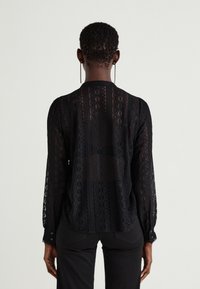 Schwarze Spitzenbluse mit langen Ärmeln, die ein transparentes Design, filigrane Muster und mit Knöpfen verschlossene Manschetten aufweist. Rückansicht, die den strukturierten Stoff zeigt.