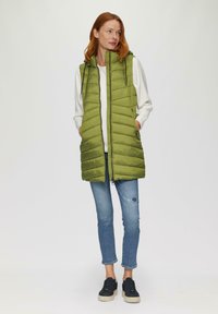 Grüne gepolsterte Weste mit Kapuze, ausgestattet mit vertikalen Nähten und zwei Seitentaschen, kombiniert mit hellblauen Jeans und schwarzen Sneakers.