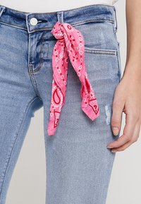 Tela de bandana rosa atada en un nudo en la cintura de unos jeans de mezclilla azul claro desgastados. La bandana presenta un patrón de paisley.