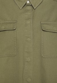 Oliwkowa zielona koszula z teksturowanego materiału, z zapinanym przodem, spiczastym kołnierzykiem i dwiema kieszeniami na piersi. Wykończona kontrastowym szyciem i perłowym guzikiem.