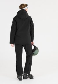 Schwarze Skijacke mit Kapuze, kombiniert mit schwarzen Schneehosen. Die Person hält einen grünen Skihelm. Fokus auf funktionales Design und Materialien.
