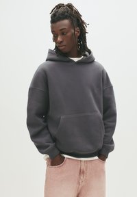 Mørkegrå hætte sweatshirt med en frontlomme, ribbede ærmer og kant. Båret over en hvid skjorte og parret med lyse pink jeans.