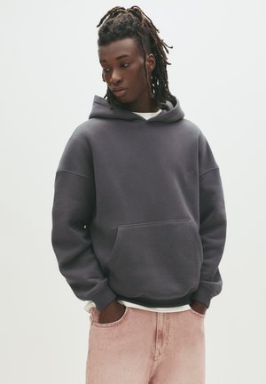 Dark grey hooded sweatshirt with a front pocket, ribbed cuffs, and hem. Worn over a white shirt and paired with light pink jeans.