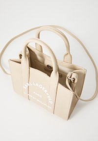 Beige läderhandväska med texturerad yta, två handtag upptill och en avtagbar axelrem. Har vit tryckt varumärkesnamn och adress.