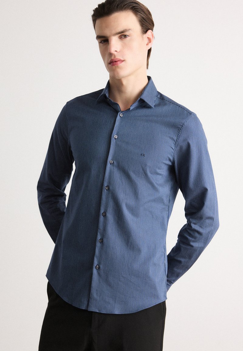Cămașă albastră cu dungi, cu mâneci lungi, guler cu nasturi, având nasturi închisi la culoare și un design slim fit. Textură netedă, stil formal.