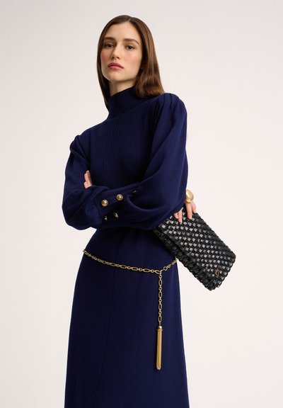 Marineblauwe geribbelde trui met pofmouwen en gouden knopen, gecombineerd met een zwarte gevlochten clutch en een gouden kettingriem met een kwastaccent.