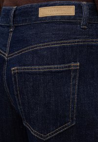 Σκούρα μπλε τζιν από denim με εμφανείς κίτρινες ραφές και πίσω τσέπη, με μια καφέ δερμάτινη ετικέτα με την ένδειξη "VERO MODA" στη μέση.