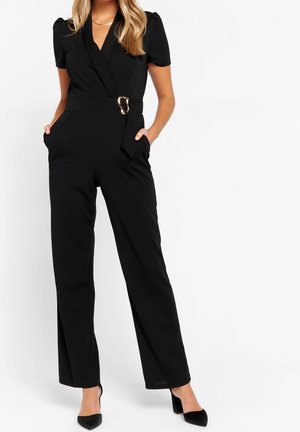 Femme portant une combinaison noire à manches courtes ceinturée avec jambes larges et talons noirs à bouts pointus, debout les mains dans les poches.