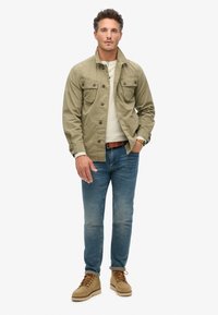 Khaki lichtgewicht jack met knoopzakken, gedragen over een wit shirt met lange mouwen, gecombineerd met blauwe jeans en beige schoenen.