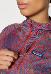 Chaqueta ligera de diseño en rojo y azul con un collar alto, bolsillo con cremallera y un logotipo negro de Patagonia en el pecho. Textura suave.