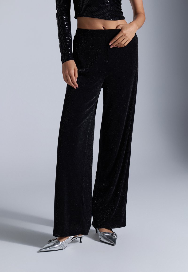 Top court en sequins noirs associé à un pantalon large noir, avec un toucher doux et un léger éclat. Modèle portant des chaussures métalliques à pointe.
