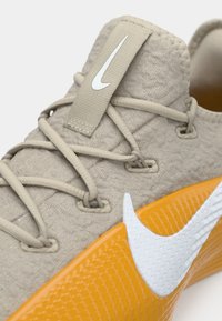 Chaussures Nike avec une tige beige texturée, des lacets gris et une semelle en caoutchouc orange arborant un logo swoosh blanc.