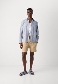 Stribet blå og hvid skjorte med knaplukning over en hvid tanktop, parret med beige shorts og blå slidesandaler. Enkel, afslappet design.