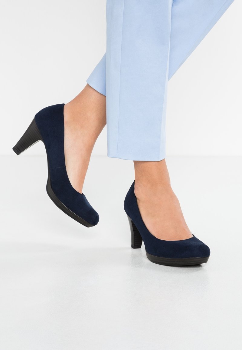 Jambes portant un pantalon bleu clair et des escarpins bleu marine en daim à talon moyen sur un sol blanc, un pied légèrement relevé derrière l'autre.