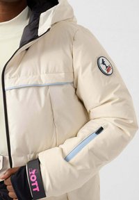 Giacca imbottita beige con cappuccio, dettagli neri, tasca laterale con cerniera e toppa rotonda con logo. Presenta una texture liscia e isolante.