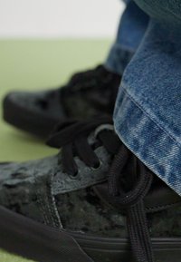 Adidași negri cu un finisaj din catifea texturată, cu șireturi negre și talpă din cauciuc, asortați cu blugi albaștri uzați.