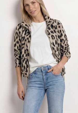 Kvinde iført en beige og sort leopardmønstret lynlåstrøje, hvid t-shirt og lyseblå jeans, stående med den ene hånd i lommen.