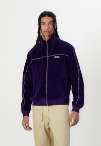 Lilla velour zip-jakke med sidelommer, kontrasterende hvit piping og brodert "JACKER" logo på brystet. Glatt tekstur.