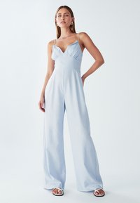 Mujer vistiendo un mono de pierna ancha de color azul claro con tirantes finos y sandalias blancas, de pie frente a un fondo blanco liso.