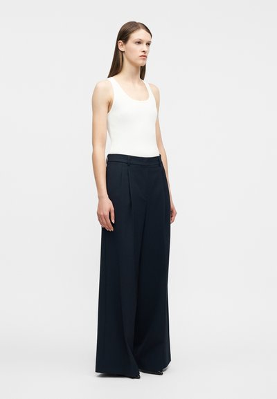 Γυναίκα που φοράει λευκό ριμπ αμάνικο τοπ και ψηλόμεσα, φαρδιά μπλε-σκού ρα παντελόνια, στέκεται μπροστά σε λευκό φωτεινό φόντο.