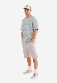 Pruhované tričko s krátkým rukávem v jemné modré a bílé barvě, spárované se světle béžovými šortkami. Model má na sobě šedou baseballovou čepici a světlé tenisky.