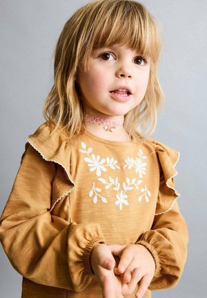 Haut marron à manches longues avec poignets à volants et broderie florale blanche. L'enfant porte un collier ras du cou rose. Fond simple, tons neutres.