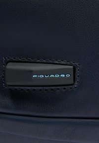Marineblauw leer met een gestructureerde afwerking, voorzien van een zwarte accentlap gestikt langs de rand, met "PIQUADRO" in lichtblauwe letters.