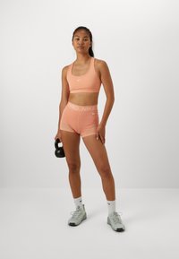 Peach sports bra and shorts set with white Nike logo, short cut, complemented by light gray sneakers and black kettlebell.