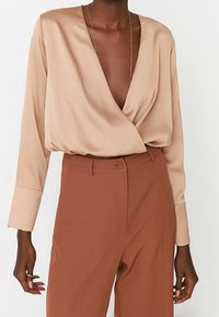 Beige indpakningsbluse med lange ærmer, blødt stof og dyb V-udskæring. Kombinationen med høj talje, rustfarvede bukser og diskrete hardware detaljer.