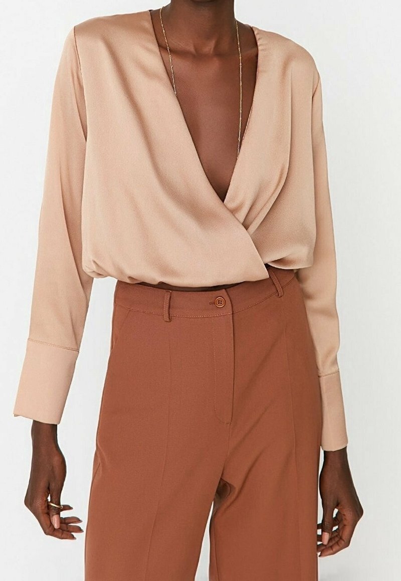 Beige indpakningsbluse med lange ærmer, blødt stof og dyb V-udskæring. Kombinationen med høj talje, rustfarvede bukser og diskrete hardware detaljer.