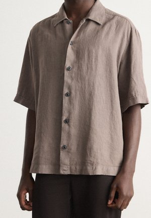 Man wearing a loose-fitting, short-sleeve beige button-up shirt with a collar and dark pants against a plain background.