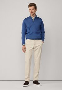 Pull à col zippé bleu à manches longues, associé à un chino beige et des baskets noires. La tenue présente un détail de col intérieur rayé.