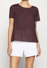 Korte mouwen, bordeauxrood top met witte stippen, ronde halslijn en borstzakje. Gecombineerd met witte shorts voor een casual look.