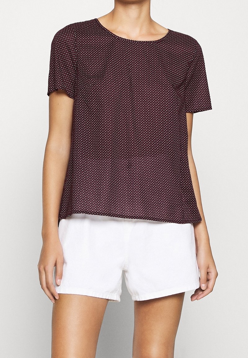 Korte mouwen, bordeauxrood top met witte stippen, ronde halslijn en borstzakje. Gecombineerd met witte shorts voor een casual look.