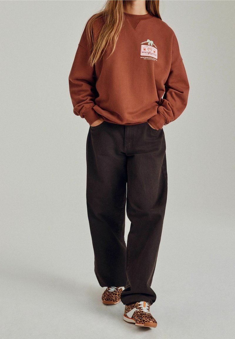 Personne portant un sweat marron avec un motif d'épices hivernales, un pantalon noir ample et des baskets à imprimé léopard, debout les mains dans les poches.