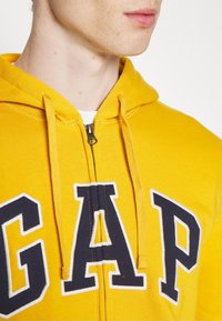 Sudadera con capucha amarilla con cremallera, con letras "GAP" en azul marino, capucha con cordón y textura suave. Detalle de cremallera en el centro.