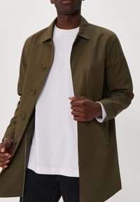 Olajzöld vízálló dzseki galléros nyakkal és gombos záródással. A sima textúra és a kontrasztos fehér varrás jellemzi.