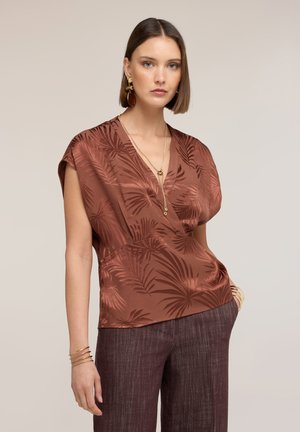 Frau mit kurzem braunem Haar, die eine rostbraune Satinbluse mit Blattmustern, braune Hosen, goldenen Schmuck und Armbänder trägt und mit einer Hand in der Tasche steht.