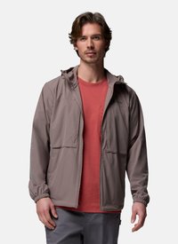 Homme portant une veste taupe zippée sur une chemise rouge et un pantalon gris, debout devant un fond clair uni.