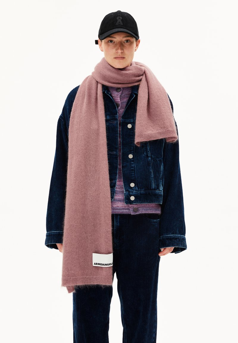 Giovane con un cappello nero, giacca di velluto a coste blu scuro oversize e pantaloni, maglione viola chiaro e lungo sciarpa rosa avvolta attorno al collo.