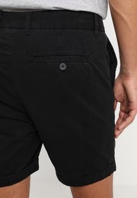 Homme portant un short noir avec des poches arrière à boutons et un t-shirt gris, visible de la taille jusqu'en bas sur le côté droit.
