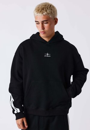 Jeune personne aux cheveux blonds courts portant un sweat à capuche noir avec un logo blanc et des rayures sur les manches, regardant vers le bas avec une main dans la poche.