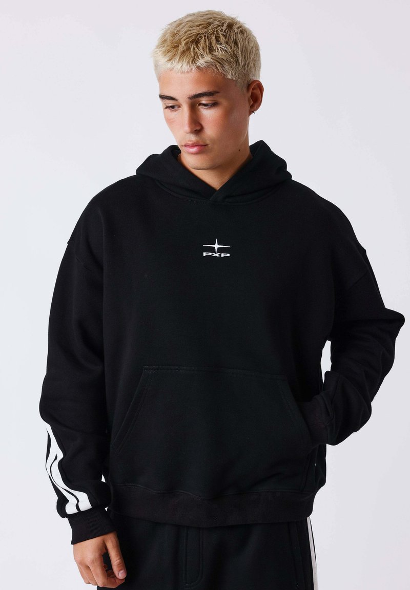 Jeune personne aux cheveux blonds courts portant un sweat à capuche noir avec un logo blanc et des rayures sur les manches, regardant vers le bas avec une main dans la poche.