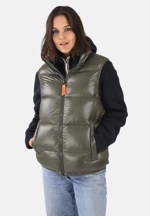 Gilet rembourré vert avec une texture brillante, fermeture éclair sur le devant, deux poches latérales et une étiquette d'accent orange, porté sur un sweat à capuche noir.